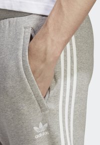 Šedé tepláky s bílými pruhy a logem Adidas. Měkká textura, boční kapsy a elastický pas. Pohodový design.