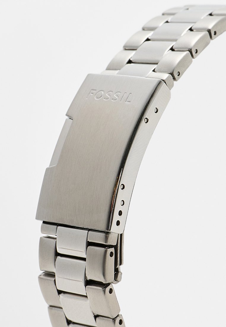 Montre en acier inoxydable argenté avec une finition brossée et un bracelet à maillons. Comprend une fermoir avec le logo "FOSSIL" et des crans réglables.