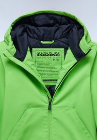 Giacca verde brillante con chiusura a zip, fodera interna nera e un'etichetta appesa in evidenza che descrive la sua costruzione in materiali sostenibili.