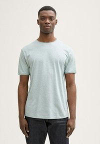 Camiseta de manga corta verde claro con escote redondo, textura suave y un detalle de logo sutil en el pecho, combinada con jeans oscuros.