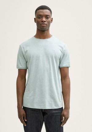 Camiseta de manga corta verde claro con escote redondo, textura suave y un detalle de logo sutil en el pecho, combinada con jeans oscuros.