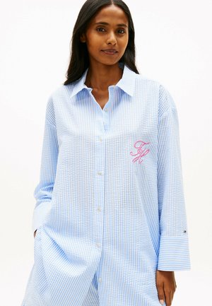 Mujer vistiendo una camisa holgada a rayas azul claro y blanca con iniciales bordadas en rosa en el pecho y con las manos en los bolsillos.