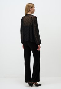 Schwarze durchsichtige Bluse mit strukturierten Polka-Dots, langen Ärmeln und lockerer Passform, kombiniert mit schwarzen weit geschnittenen Hosen und schwarzen High Heels.