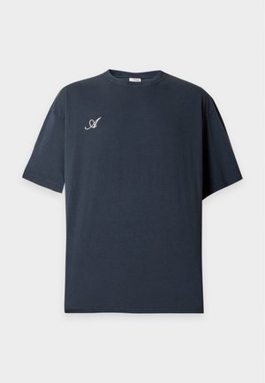Muž v čiernom tričku Arkk Copenhagen s bielym textom na chrbte, v modrých džínsoch, stojaci pred jednoduchým bielym pozadím.