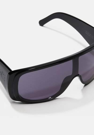 Óculos de sol pretos com uma armação larga e lentes cinza-escuras. Apresentam um acabamento suave e uma ligeira curva ao longo da borda superior da armação.