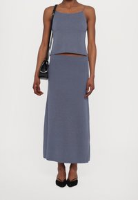 Two-piece set in soft, ribbed fabric. Gray color, sleeveless cropped top with thin straps and a high-waisted, floor-length skirt. Black handbag.
