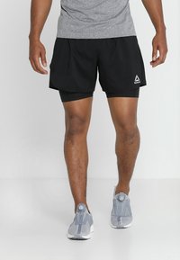 Černé sportovní šortky s integrovanými legínami, vyrobené z lehkého materiálu, s bílým logem Reebok a bočními kapsami. Šedé tenisky dotvářejí celkový vzhled.