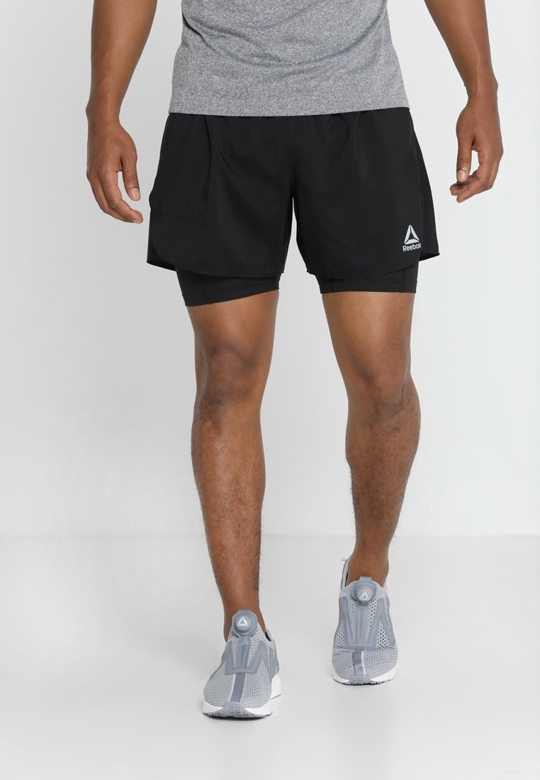 Černé sportovní šortky s integrovanými legínami, vyrobené z lehkého materiálu, s bílým logem Reebok a bočními kapsami. Šedé tenisky dotvářejí celkový vzhled.