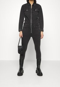 Combinaison en denim noir avec fermeture éclair à l'avant, deux poches poitrine, coupe ajustée et jambes fuselées. Associée à des bottes noires épaisses et un petit sac.