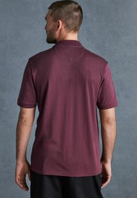 Polo majica kratkih rukava u burgundskom s tonu teksturiranog uzorka. Ima klasičan ovratnik i ležeran kroj, prikazana s leđa.
