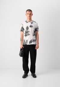 Witte t-shirt met zwart tropisch bladerenpatroon, gecombineerd met zwarte broek en zwarte schoenen. Houdt een kleine zwarte tas vast. Minimalistische achtergrond.