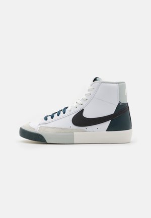 Baskets montantes blanches avec le swoosh Nike noir, des touches gris et vert foncé, des lacets blancs et une semelle plate sur un fond uni.