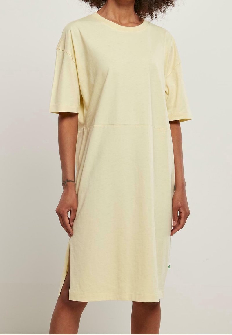 Femme portant une robe ample, jaune pâle, à manches courtes et longueur genou avec une fente sur le côté, posant devant un fond uni.
