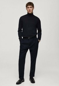 Schwarzer Rollkragenpullover kombiniert mit dunklen Hosen. Glatte Textur, taillierte Passform und Lederschuhe runden das Outfit ab.