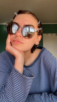 Óculos de sol redondos com lentes refletoras, detalhes em plástico transparente, usados com uma camisa de manga longa às riscas. Tom de pele claro, cabelo curto com presilhas.