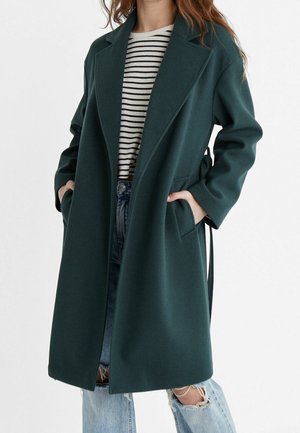 Femme portant un long manteau vert foncé sur un haut rayé noir et blanc et un jean déchiré bleu clair, les mains dans les poches du manteau.