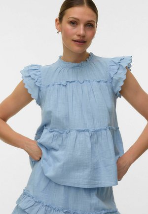 Femme portant un chemisier sans manches bleu clair avec des manches à volants et des détails à volants en étages, posant avec les mains sur les hanches.