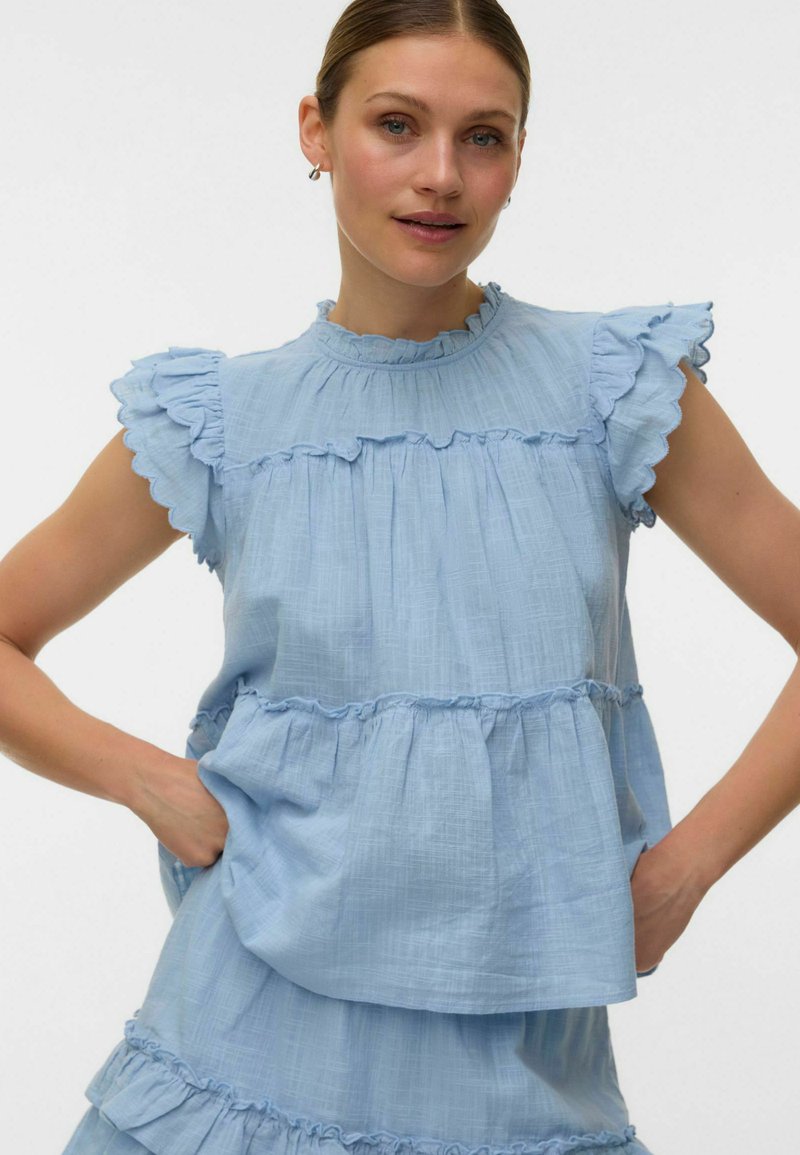 Femme portant un chemisier sans manches bleu clair avec des manches à volants et des détails à volants en étages, posant avec les mains sur les hanches.