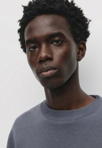 Grå sweatshirt med ribbet rund hals og struktureret stof, med et nærbillede af en models ansigt og mørkt, krøllet hår mod en lys baggrund.
