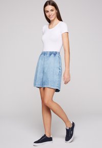 Falda de mezclilla azul con cintura elástica, hasta la rodilla, que presenta una textura suave y costuras laterales. Combinada con una camiseta blanca ajustada y zapatillas.