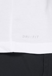 Camiseta Dri-FIT blanca con una textura suave; presenta un logo impreso sutil en gris cerca del dobladillo. Combinada con un tejido negro por debajo.