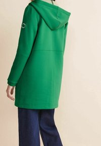 Felpa verde con cappuccio dalla texture liscia, con un logo circolare sulla manica, abbinata a jeans a gamba larga di colore blu scuro.