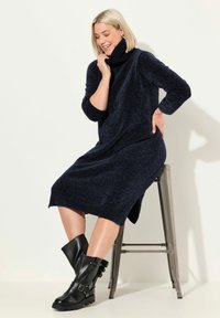 Abito lavorato a maglia blu navy con colletto alto, maniche lunghe e spacchi sui lati. Indossato con stivaletti neri in pelle con fibbie decorative.
