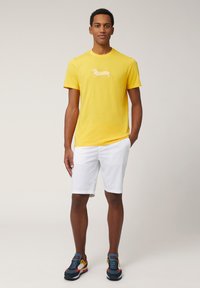 Gele katoenen t-shirt met een gestreepte teckelafbeelding, gecombineerd met witte shorts en multicolore sport sneakers.