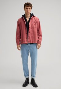 Lichtroze oversized shirtjacket met knoopsluiting, borstzak, gelaagd over een zwarte hoodie, gecombineerd met opgerolde lichtblauwe jeans en zwarte laarzen.