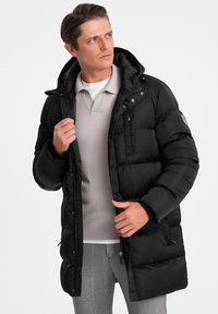 Giacca a vento nera con zip frontale, cappuccio e tessuto isolante. Presenta un design trapuntato e tasche laterali, indossata sopra una polo grigia chiara.