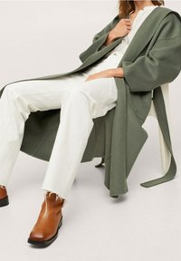 Green wool blend coat with wide sleeves, worn over a white shirt and distressed white pants, complemented by brown ankle boots.
