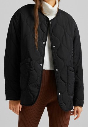 Vrouw draagt een zwarte gewatteerde jas met drukknopen aan de voorkant over een witte coltrui en bruine geribbelde broek tegen een grijze achtergrond.