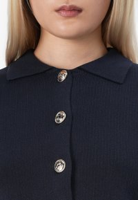 Vrouw draagt een marineblauwe gebreide cardigan met een kraag en drie decoratieve gouden knopen aan de voorkant.