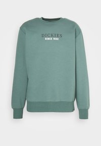 Grön sweatshirt med rundad hals, ribbade ärmslut och nederkant. Har en tryckt logotyp, "DICKIES SINCE 1922," i mörka bokstäver på framsidan.