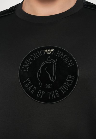 Juodi marškinėliai su apvalia aksomine aplikacija su arklio atvaizdu, įspaustu pilku užrašu „EMPORIO ARMANI“ ir „YEAR OF THE HORSE“.