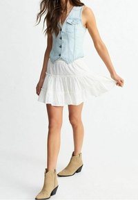 Veste en denim bleu clair avec boutons, portée sur une robe blanche en dentelle à volants. Bottines camel avec ornements complètent la tenue.