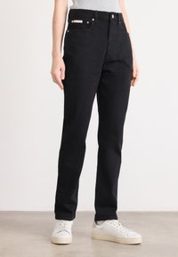 Persona che indossa jeans neri a gamba dritta con sneakers bianche su un pavimento grigio chiaro e sfondo bianco.