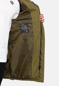 Olijfgroene winterjack met ritssluiting, gevoerde binnenkant en een etiket. Beschikt over een binnenzak en zijknoppen voor extra functionaliteit.