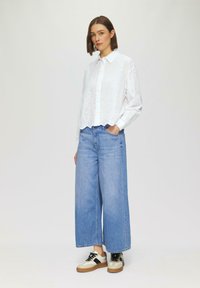 Witte oogjes knoopshirt met lange mouwen, gecombineerd met lichtblauwe wijde jeans en meerkleurige sneakers met zwarte veters.