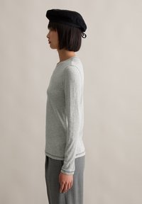 Hellgraues Langarmshirt mit rundem Halsausschnitt, aus weichem Material. Kombiniert mit grauen, plissierten Hosen und einer schwarzen Baskenmütze.