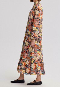 Robe maxi florale avec des motifs vibrants orange, jaune et bleu. Manches longues, col monté et ourlet volanté. Des chaussures plates noires complètent le look.