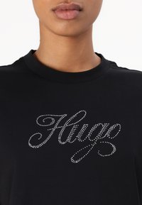 Black cotton t-shirt featuring a silver rhinestone logo design that spells "Hugo" in a cursive font, round neckline, and short sleeves.