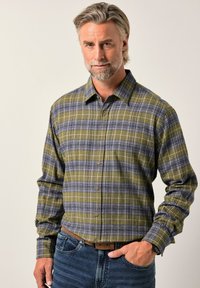 Středně starý muž s šedým vousem, oblečený v kostkované košili s zelenými a šedými pruhy, modrých džínách a hnědým opaskem, stojící s jednou rukou v kapse.