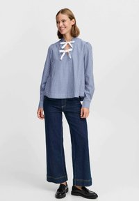 Blouse longue à rayures bleues et blanches avec un décolleté en trou de serrure, des volants, et des accents en ruban blanc, associée à un jean noir à jambes larges et des chaussures noires.