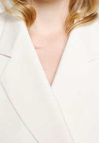 Biała, dopasowana marynarka z głębokim dekoltem w serek, ostrymi klapami i gładką fakturą. Posiada subtelne przeszycia w pobliżu kołnierza.