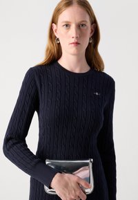 Tamsiai mėlynas pynimo raštu megztinis su ilgomis rankovėmis, apvaliu iškirpimu ir mažu siuvinėtų logotipu, derinamas su metalizuoto sidabro rankine.