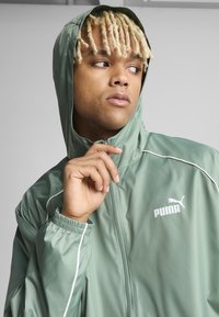 Grønn lett jakke med hette, med en hvit Puma-logo og kontrasterende hvit piping langs glidelåsen og ermene. Jevn tekstur.