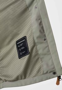 Innenansicht einer olivgrünen Jacke mit atmungsaktivem Netzfutter, Druckknöpfen, Reißverschluss mit Lederlasche und Swedemount-Label mit wasserdichten und winddichten Symbolen.