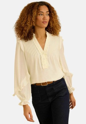 Bílá halenka s plisovaným předním dílem a volánkovými manžetami, kombinovaná s tmavými džínovými kalhotami a hnědým páskem. Hladká textura, měkká látka.