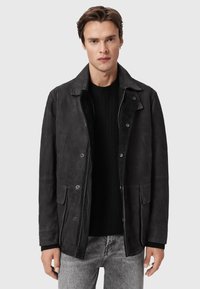 Homme portant une veste en suède noire avec des poches à rabat sur un pull noir et un jean gris clair, se tenant contre un fond clair et uni.